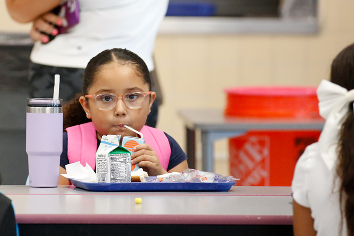 A little girl in glasses sips a juice box in the cafeteria