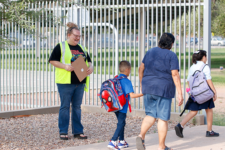 A woman in a yellow safety vest smiles as a boy and girl and their mom walk in to the school courtyard