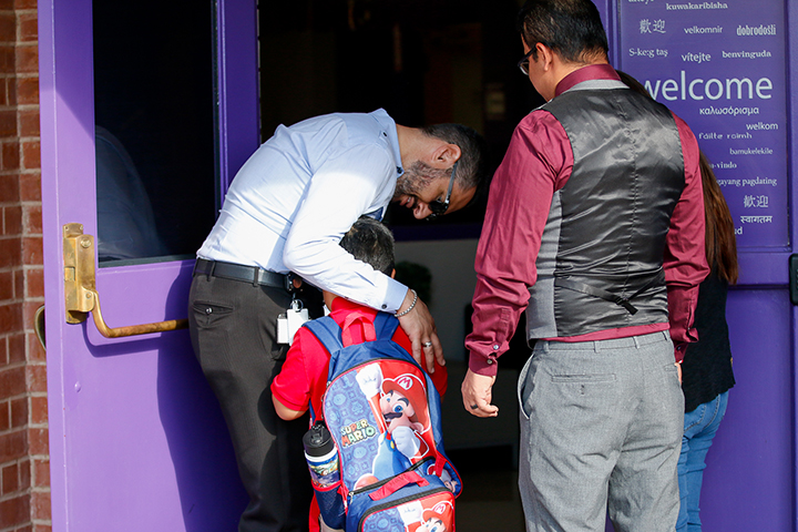 A man with a beard and sunglasses hugs a young boy as his father walks him into school