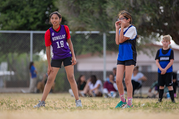 A girl in a Bonillas jersey and a girl in a Dunham jersey stand on the field